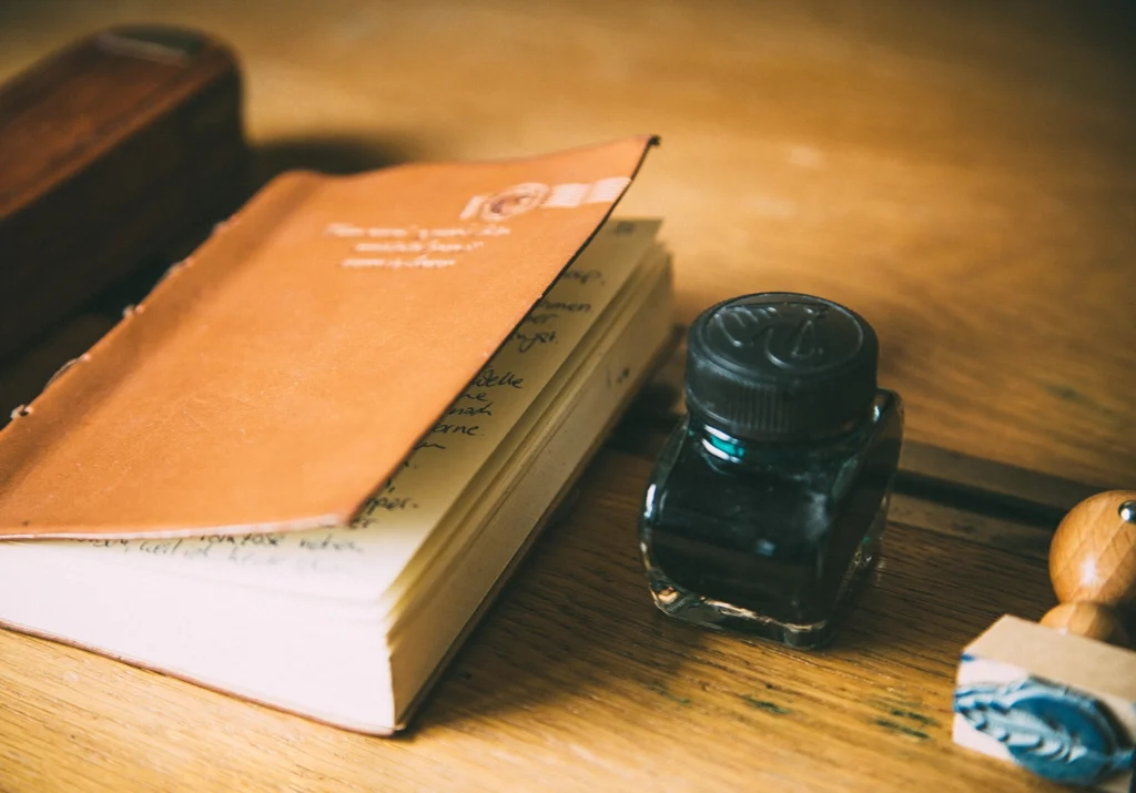 Ein Notizbuch, ein Tintenfass und ein Stempel mit dem Logo von aufs-papier liegen auf einem Holztisch