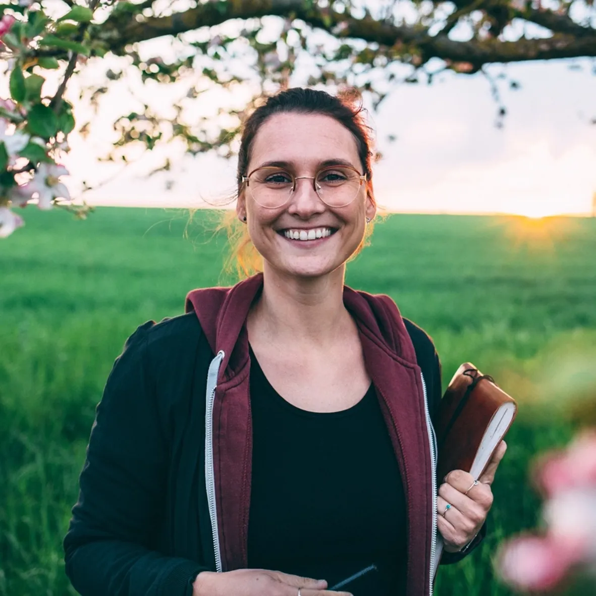 Irena steht mit Stift und Block in der Hand lächelnd in die Kamera schauend vor einem Sonnenuntergang an einer grünen Wiese